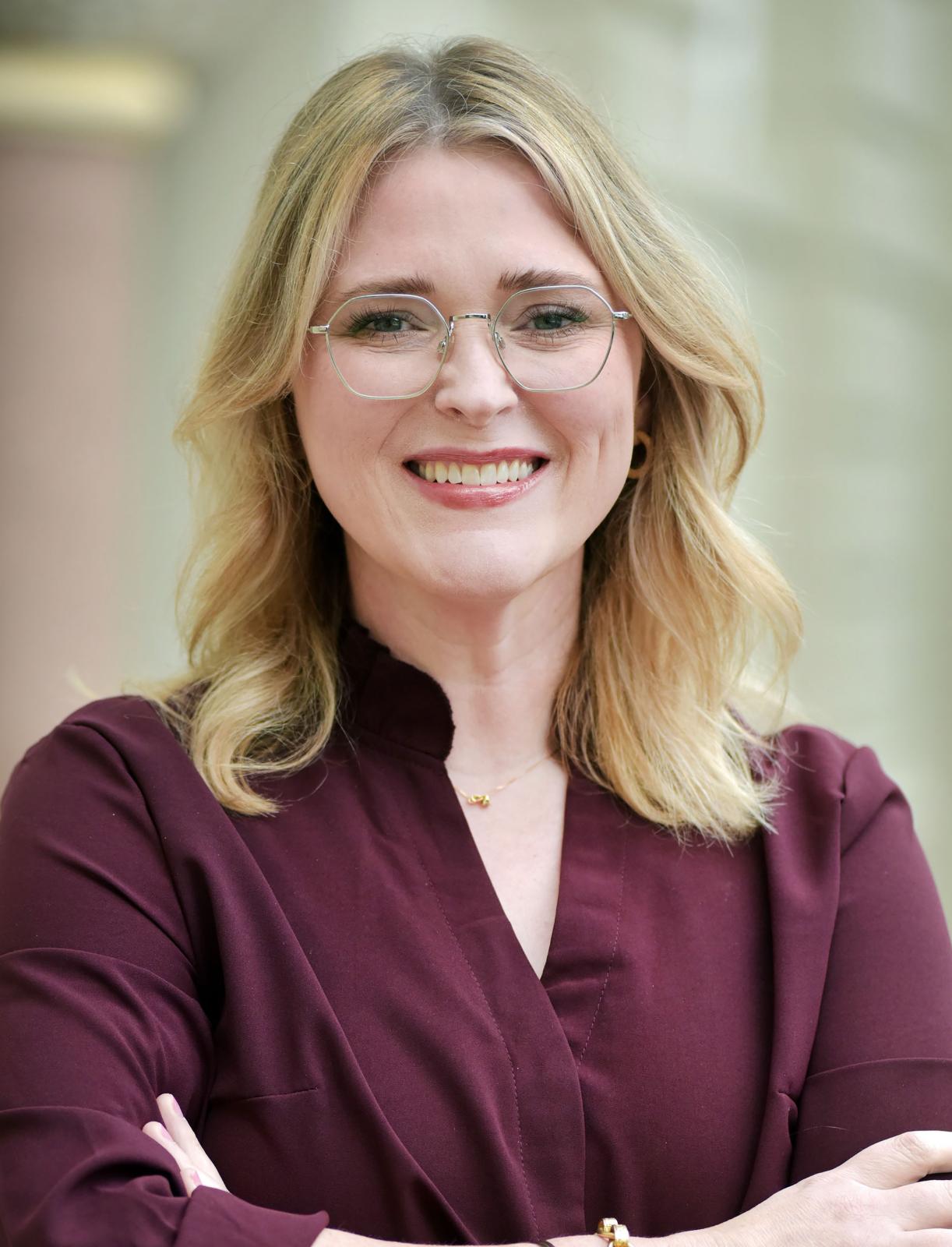 Portrait of Kayla Carr with short blonde hair and glasses wearing a maroon shirt with her arms crossed.